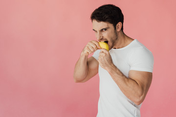 hungry muscular man in white t-shirt eating banana isolated on pink