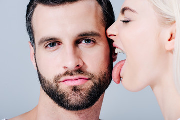 young woman licking face of man looking at camera isolated on grey