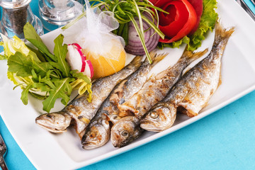 Fototapeta na wymiar Mediterranean dish, European cuisine. Whole fish grilled capelin, served with a salad of vegetables — greens, arugula, onion rings, tomato, green and yellow pepper and lemon
