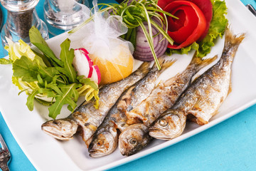 Mediterranean dish, European cuisine. Whole fish grilled capelin, served with a salad of vegetables — greens, arugula, onion rings, tomato, green and yellow pepper and lemon