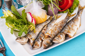 Mediterranean dish, European cuisine. Whole fish grilled capelin, served with a salad of vegetables — greens, arugula, onion rings, tomato, green and yellow pepper and lemon