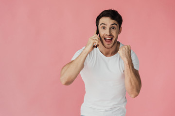 front view of excited muscular man in white t-shirt talking on smartphone and showing yes gesture isolated on pink