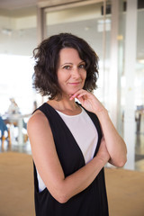 Pensive smiling business woman posing in office building hall. Middle aged woman in casual standing with arms folded, touching chin and smiling at camera. Female portrait concept