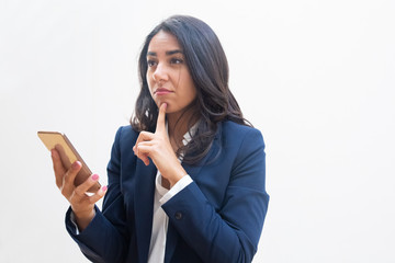 Thoughtful young woman with smartphone. Beautiful young businesswoman standing with finger on chin...