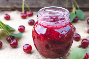 Bank with homemade cherry jam on a wooden background near the berries and leaves.