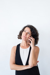 Pensive woman talking on cellphone in studio. Middle aged curly haired lady in casual standing over white background and calling on mobile phone. Phone conversation concept