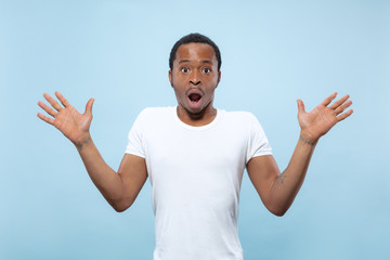 Half-length close up portrait of young african-american man in white shirt on blue background. Human emotions, facial expression, ad, sales concept. Inviting, looks shocked and astonished. Copyspace.