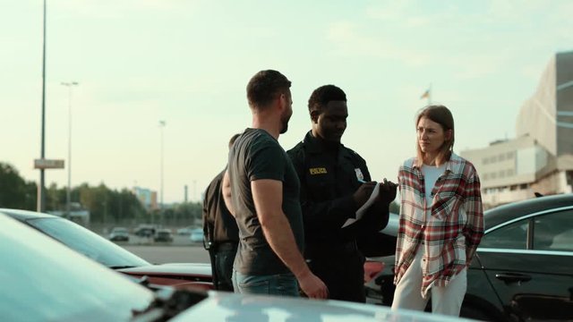 Policeman afro-american on duty talking with young stressed people at road accident area. Two upset drivers argue try to solve the situation. Police patrol.
