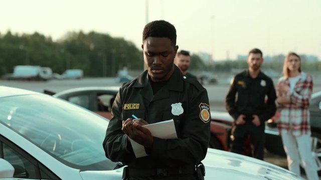 Focused policeman afro-american writing notes on papers describing a road accident in detail. Portrait of handsome cop against people drivers and policeman argue stand near the crashed car.