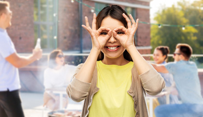 people, gesture and portrait concept - smiling young asian woman looking through finger glasses over rooftop party background