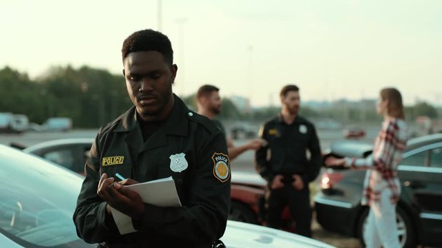Portrait of happy african policeman doing his job with enjoy smiling broadly leaning on police car. In background man and woman drivers argue with policeman at car crash accident area.