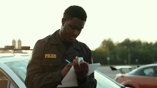 Portrait of young black police officer writing notes at accident investigation. Close-up afro cop doing paperwork turning on the car crash scene with police on background.