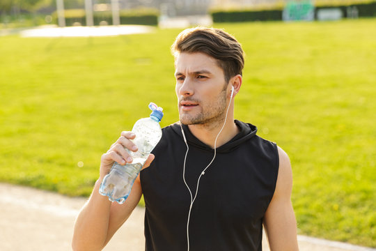 Image of masculine man drinking water while working out with earphones and running at green park outdoors in morning