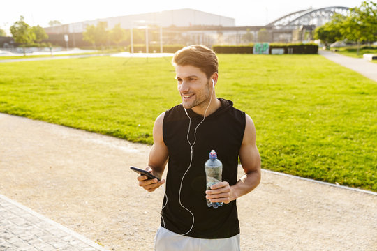 Image of muscular man drinking water and holding cellphone while running with earphones at green park outdoors