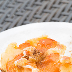 Delicious apricot cake with powdered sugar and cinnamon on a white plate. In the blurred background, on a dark grey rattan table, there is copy space.