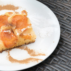 Delicious homemade apricot cake on a white tableware. On the white plate are the cinnamon and sugar masks of a spoon and a fork visible. Dark grey rattan table in the background.