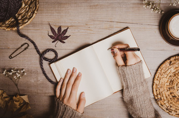 Fall winter background. Female hand holding a pen for a sketch or letter on old notebook with a ball of wool yarn and cup of cappuccino on a wooden background. Home hobbies, Cozy comfort concept.