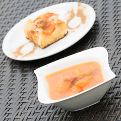 In the front tasty apricot puree with cinnamon in a white dishware. In the background there is an apricot cake with cinnamon and powdered sugar visible.