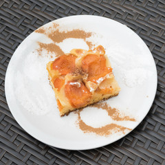 Fruit flan with a white plate on a grey table. On the white plate there are is a mask of  two forks and two spoons with cinnamon and powdered sugar visible. The cake is made with apricots. 
