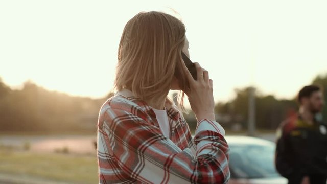 Disappointed young woman talking on the phone standing in the street at road accident. Mixed-up girl in shirt asking for help calling for the police. Car crash.