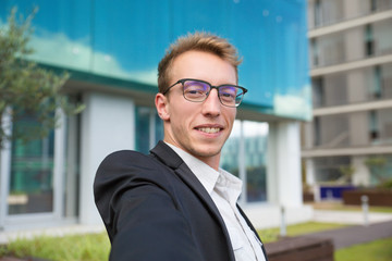Selfie of happy cheerful businessman in urban settings. Handsome young business man in eyeglasses holding smartphone in outstretched hand and showing and looking at phone camera. Photo concept