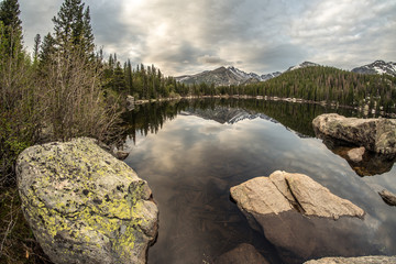 Snow covered mountains reflected and an alpine lake with mirror flat water, rocks and trees in the foreground and dramatic sky in the background, Bear Lake, Rocky Mountain National Park, Colorado
