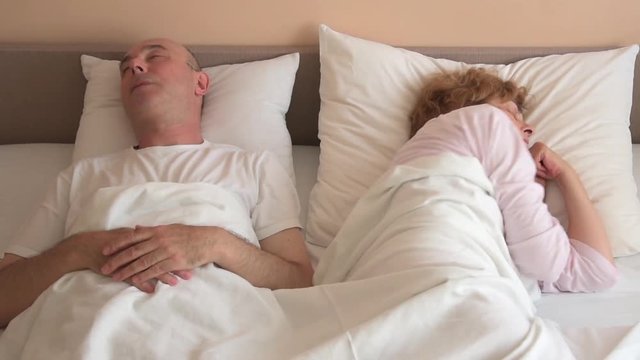Couple in bed after arguing. Man lying on his back and sleeping while angry woman looking at him and turning to the other side. 