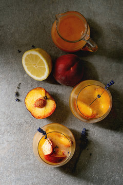 Peach cold lemonade or Bellini cocktail with lavender flowers served in two glasses with pitcher of pesh juice and lemon on dark texture background. Flat lay, space. Homemade summer drink.