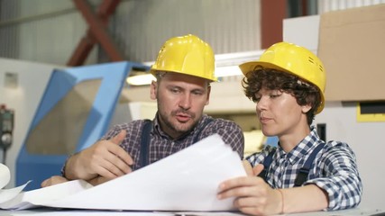 Pan chest-up shot of male and female engineers studying technical drafts and discussing manufacturing process, standing in front of sophisticated automated modern production line machinery