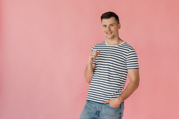 smiling man in striped t-shirt standing with hand in pocket, blinking and showing thumb up isolated on pink