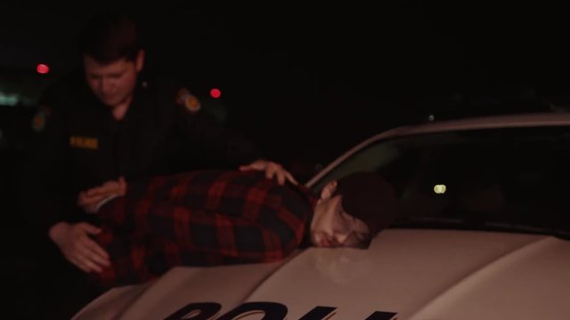 Arresting a dangerous thief on police night raid. View of young policeman searches a handcuffed drunk drug dealer on the car hood. Close-up of cop holding drugs. Crime scene.