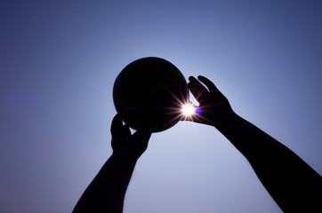 Silhouette of the ball and hands. The sun shines through the fingers. The concept of outdoor sports, summer hobby. Minimalism, cloudless sky, place for text.