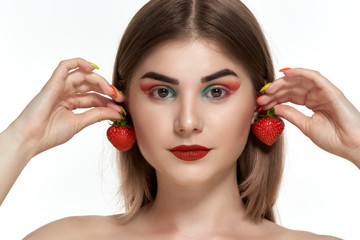 Closeup portrait of a beautiful young woman with bright color make-up holding strawberry near the face.
