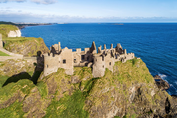 Fototapeta na wymiar Ruins of medieval Dunluce Castle on a steep cliff near Bushmills. Northern coast of County Antrim, Northern Ireland, UK. Aerial view in sunrise light. Far view of Portrush resort in the background