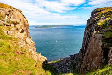 Northern Ireland, UK. Fragment of Fair Head cliff at the north-eastern corner of County Antrim with the far view of Rathlin Island, a sailboat and Atlantic Ocean