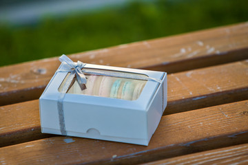 Cardboard white gift box with colorful handmade macaron cookies on wooden bench outdoors.