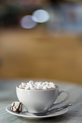 Mug of coffee with marshmallows on porcelain plate on blurred colorful interior bokeh background.