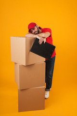 Image of tired delivery man in red uniform sleeping while standing with clipboard over packaging boxes