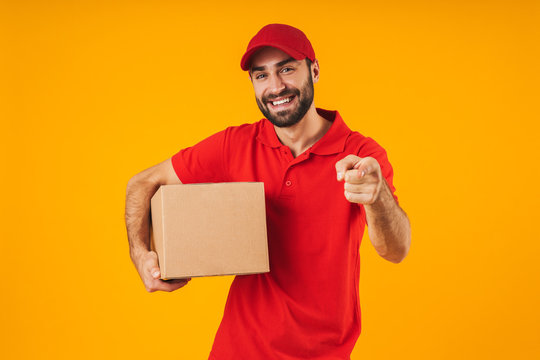 Portrait of positive delivery man in red uniform pointing finger at camera and holding packaging box