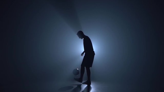 Silhouette of football freestyler at studio against blue spotlight. Slow motion