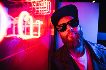 Hipster handsome man on the city streets being illuminated by neon signs. He is wearing leather...