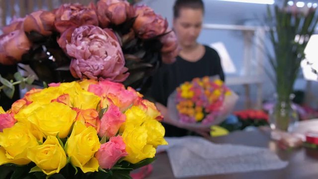 Floral business concept. Blur background with professional florist woman packaging roses boquet in flower shop. White and pink roses and peonies on table in vases. Working in floristic studio store.