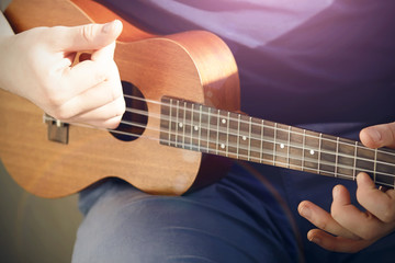 A guy in a blue t-shirt and light jeans plays a ukulele with nylon strings, illuminated by pleasant sunlight.