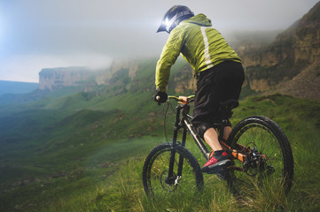 Fototapeta premium Aged male athlete in helmet and mask rides a mountain bike on a grassy slope against the background of plateau rocks and low clouds on an overcast day. Downhill mountain bike concept