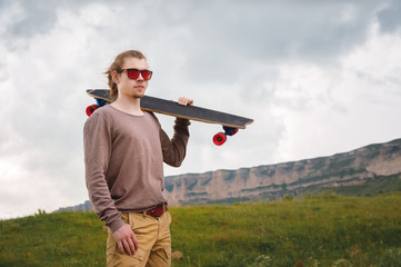 Stylish young man standing along a winding mountain road with a skate or longboard on his shoulder in the evening after sunset. The concept of youth sports and travel hobbies
