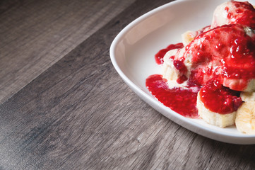 Closeup Vanilla ice cream poured with strawberry or raspberry jam with sliced banana slices on a white plate on a dark wooden table. You can do this at home on vacation