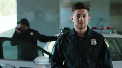 Attractive young man cops stand near patrol car look at camera around enforcement background his colleague happy officer police uniform auto safety control policeman portrait close up slow motion