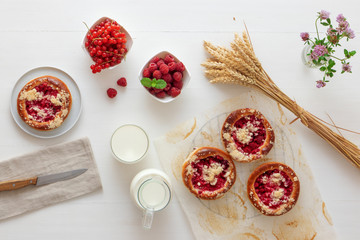 White table with yeasty sweet pastry with fresh currants and raspberries and streusel topping, glass and pitcher of milk, sheaf of wheat, small glass vase with flowers. Summer fresh fruit appetizer.