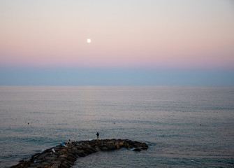 High angle seascape view with people fishing on a rocky breakwater and full moon at sunset, Diano Marina, Imperia, Liguria, Italy