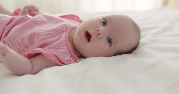 Medium shot of adorable baby girl wearing pink T-shirt lying on back in bedroom jerking legs and moving arms, periodically showing tongue and looking at operator 